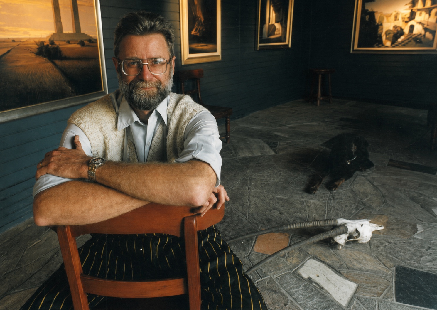 Keith Alexander sitting backwards on a wooden chair, arms folded across its back, in a dark room hung with his paintings; a black dog and an antelope skull lie on the stone floor behind him.